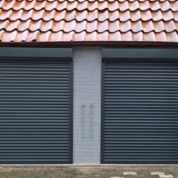 SOLNEXT Małgorzata Płonka - Dwa zamknięte, ciemnoszare systemy osłonowe na okna garażowe w jasnym, murowanym budynku z pomarańczowym dachem.