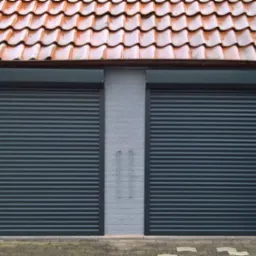 Dwa zamknięte, ciemnoszare systemy osłonowe na okna garażowe w jasnym, murowanym budynku z pomarańczowym dachem.