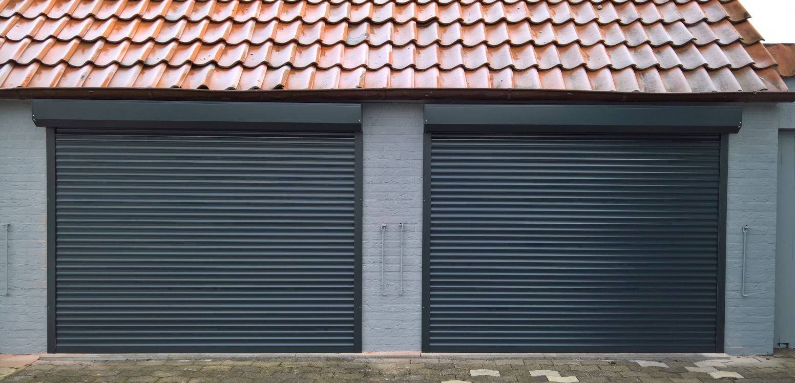 Dwa zamknięte, ciemnoszare systemy osłonowe na okna garażowe w jasnym, murowanym budynku z pomarańczowym dachem.