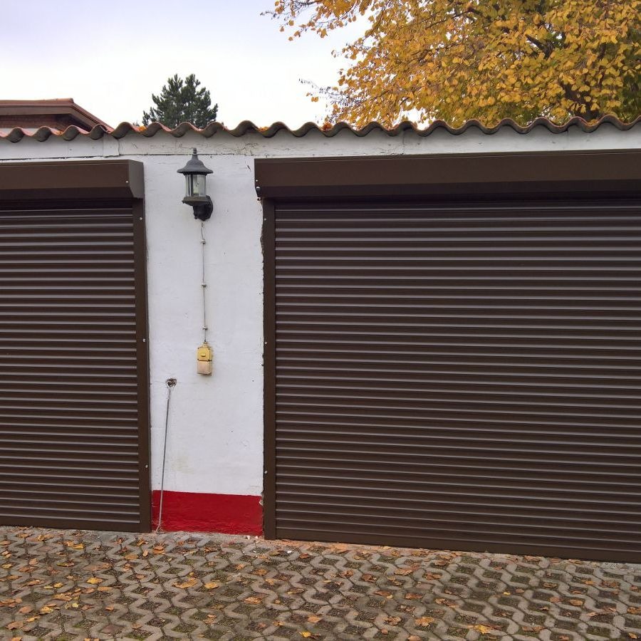 Dwa brązowe garaże z roletami zwiniętymi do połowy, oświetlone zewnętrzną lampą, na tle białej ściany z czerwoną podstawą i brukowanym podjazdem pokrytym liśćmi.