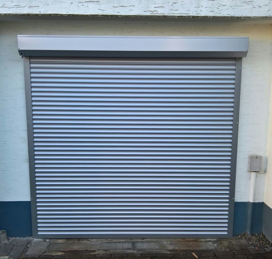 Zamknięta, szara, metalowa osłona okienna zewnętrzna z widoczną skrzynką i prowadnicami, zamontowana na elewacji budynku.