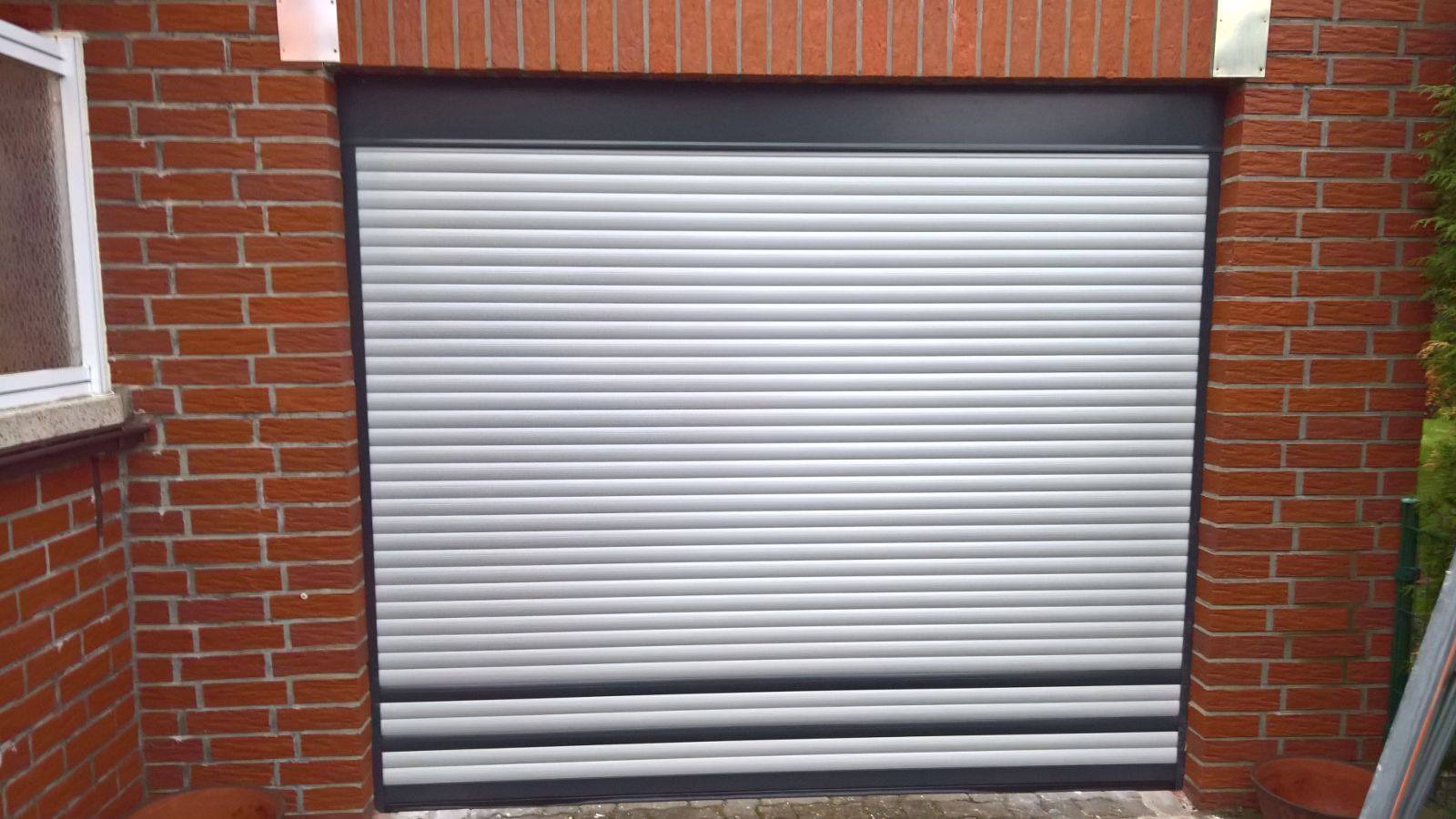 Zamknięta, aluminiowa roleta zewnętrzna w kolorze jasnoszarym, zamontowana w murze z czerwonej cegły, widoczna z zewnątrz budynku.