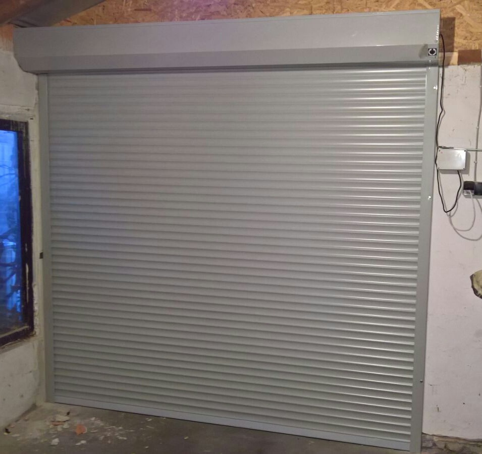 Szara roleta zewnętrzna zamontowana w surowym wnętrzu, widoczne okablowanie i drewniane elementy konstrukcyjne.