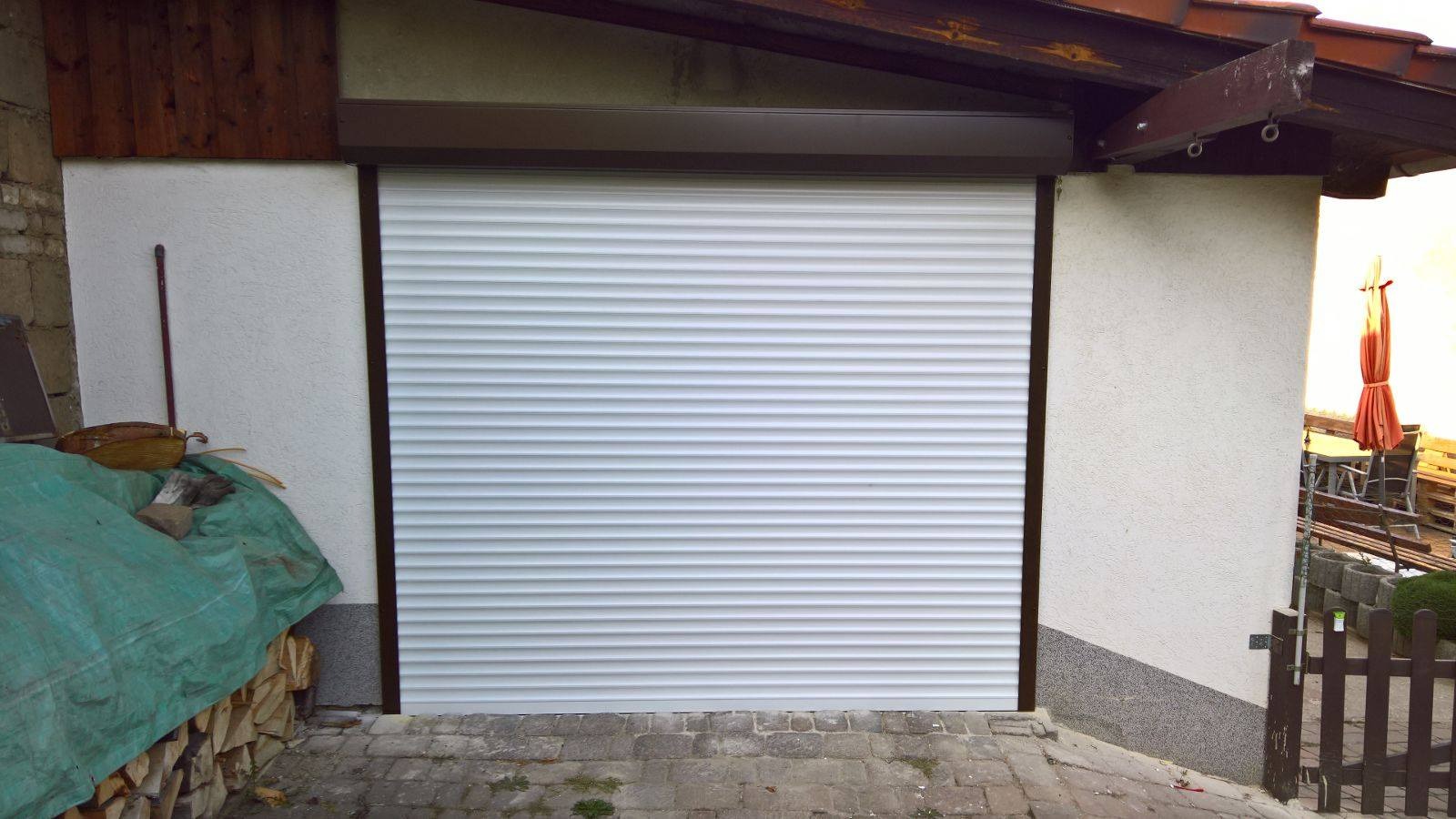 Zamknięta biała roleta zewnętrzna z brązową ramą na tle jasnej ściany budynku, widoczny stos drewna przykryty zieloną plandeką.