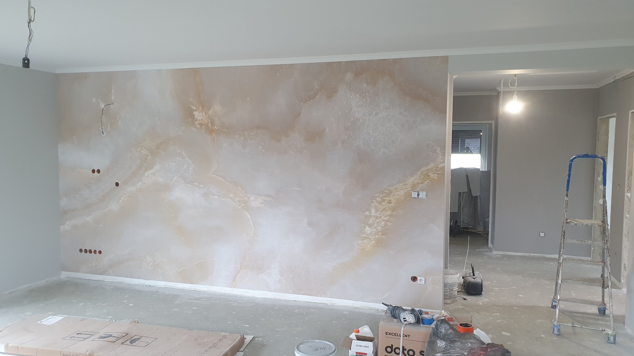 Ściana z efektem marmuru w odcieniach beżu i bieli, widoczne puszki z farbą, szlifierka i drabina w trakcie prac wykończeniowych.