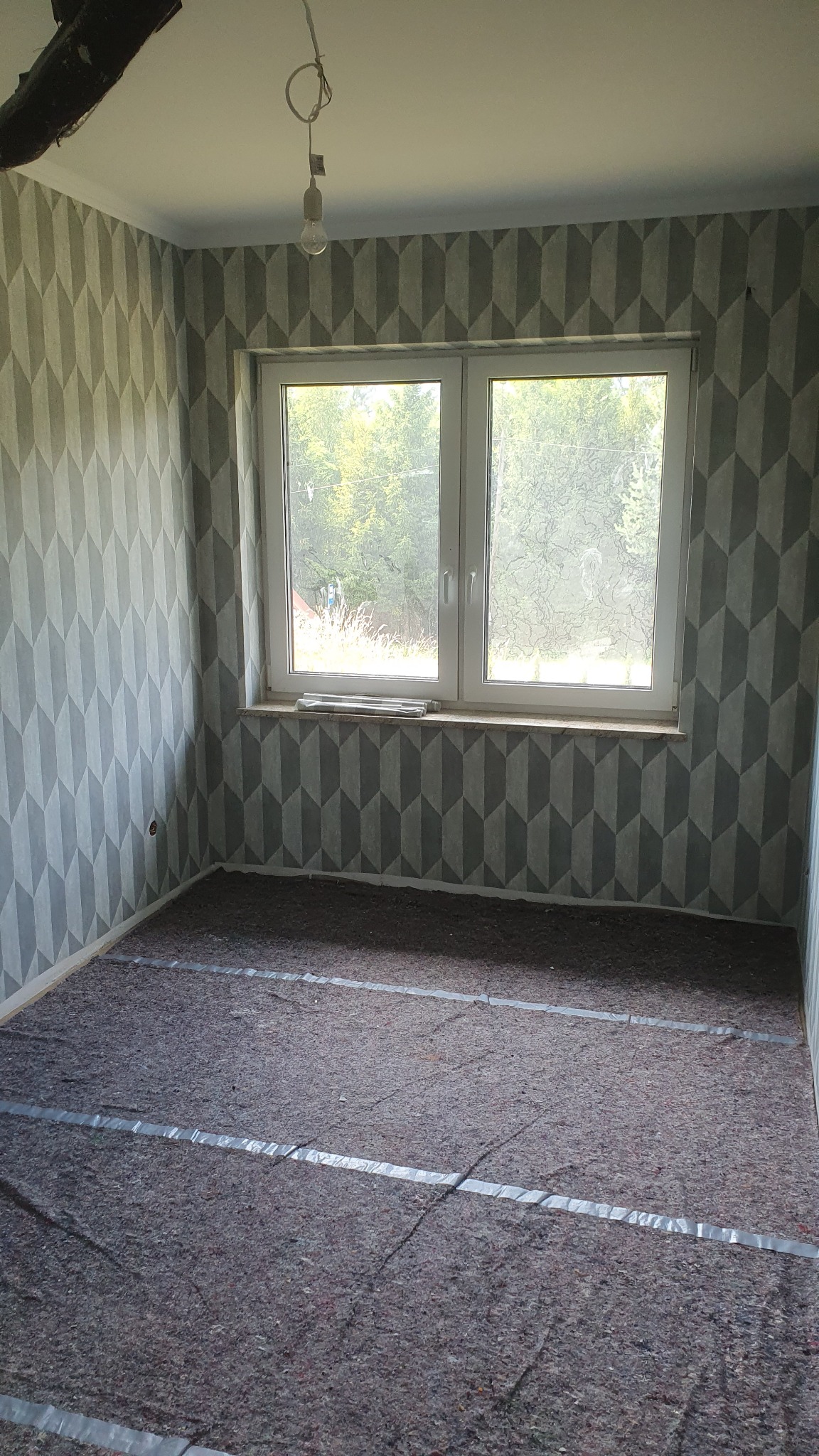 Pokój po tapetowaniu w geometryczny wzór, zabezpieczona podłoga przed układaniem paneli, surowa żarówka zwisa z sufitu.