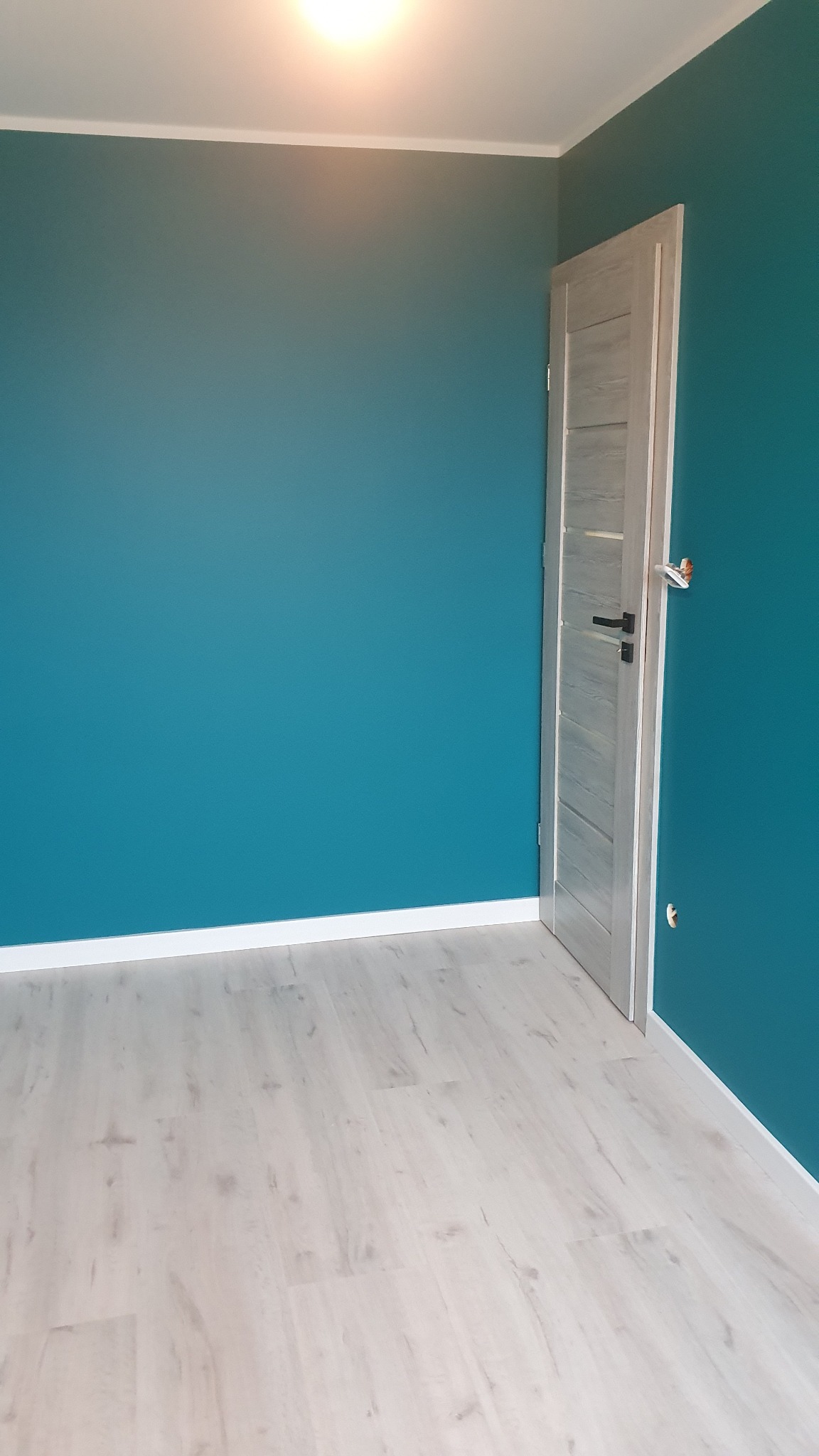 Turkusowa ściana w pokoju z jasnymi panelami podłogowymi i szarymi drzwiami z czarną klamką.