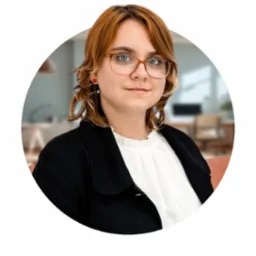 Portret kobiety w okularach, czarnym żakiecie i białej bluzce, z rozmytym tłem biurowym w okrągłej ramce.