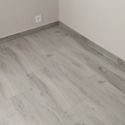 Artur Fraszczak - Ułożone jasnoszare płytki podłogowe imitujące drewno, z szarą listwą przypodłogową i gniazdkiem elektrycznym na ścianie.