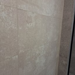 Artur Fraszczak - Ściana wyłożona beżowymi, matowymi płytkami imitującymi kamień, z delikatną strukturą i fugami w odcieniu zbliżonym do płytek, przylegająca do ciemniejszej, szarej ściany z podobną fakturą...