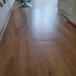 Artur Fraszczak - Ułożone brązowe panele podłogowe imitujące drewno, widoczne chromowane nogi hokerów i fragment białej szafki, realizacja w Dzierżoniowie.