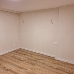 Artur Fraszczak - Pusty pokój po remoncie: nowe panele podłogowe w odcieniach brązu, świeżo pomalowane ściany w kolorze ecru, gniazdka elektryczne i wystające kable ze ściany. Widoczne białe listwy przypodłogowe...