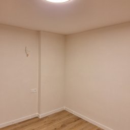 Artur Fraszczak - Pusty pokój po remoncie z panelami podłogowymi, pomalowanymi ścianami na jasny kolor i zamontowanym oświetleniem sufitowym. Widoczne wystające kable ze ściany oraz gniazdko elektryczne.