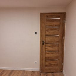 Artur Fraszczak - Wnętrze pokoju z drewnianą podłogą i drzwiami wewnętrznymi z poziomymi, metalowymi wstawkami, widoczny kontakt i włącznik na ścianie.