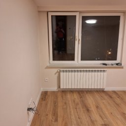 Artur Fraszczak - Pusty pokój po remoncie z drewnianą podłogą, białymi ścianami i grzejnikiem pod oknem. Na parapecie leżą narzędzia, a w oknie widać odbicie osoby robiącej zdjęcie. Ze ściany wystają kable.
