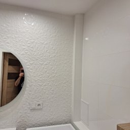 Artur Fraszczak - Biała łazienka z umywalką, pralką i okrągłym lustrem, ściany wyłożone płytkami 3D w geometryczne wzory, widoczne gniazdko elektryczne i rewizja.