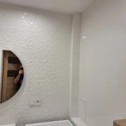 Biała łazienka z umywalką, pralką i okrągłym lustrem, ściany wyłożone płytkami 3D w geometryczne wzory, widoczne gniazdko elektryczne i rewizja.