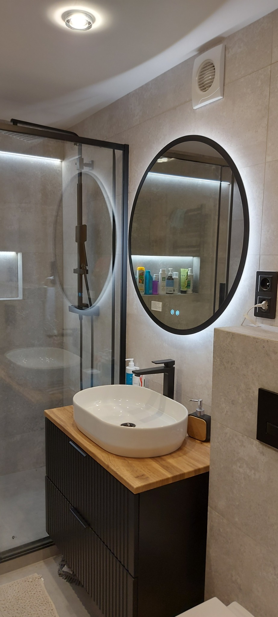 Nowoczesna łazienka z owalną umywalką nablatową, czarną armaturą i okrągłym lustrem z podświetleniem LED, widoczna kabina prysznicowa z czarnymi profilami.