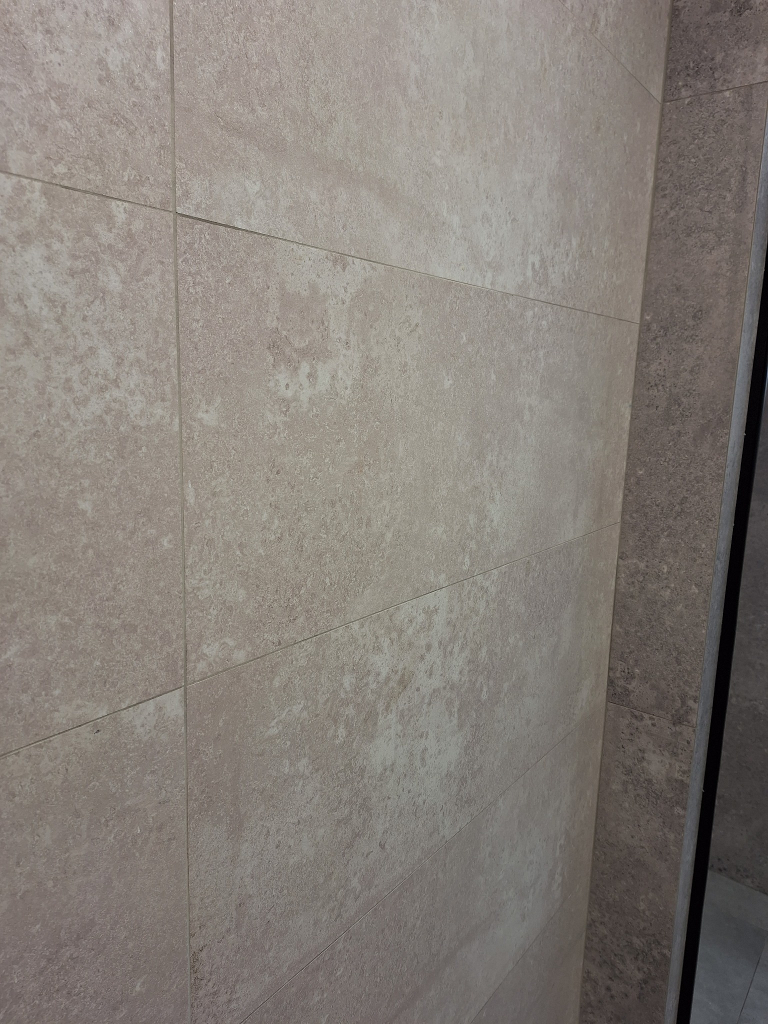 Ściana wyłożona beżowymi, matowymi płytkami imitującymi kamień, z delikatną strukturą i fugami w odcieniu zbliżonym do płytek, przylegająca do ciemniejszej, szarej ściany z podobną fakturą...