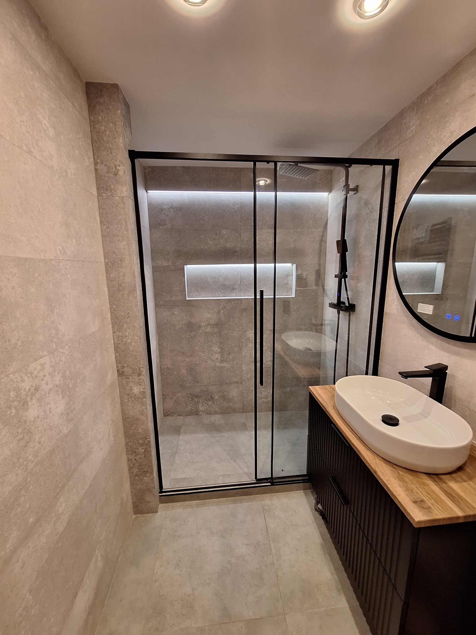 Nowoczesna łazienka z kabiną prysznicową typu walk-in z czarnymi profilami, owalną umywalką nablatową na drewnianym blacie i okrągłym lustrem z podświetleniem LED.