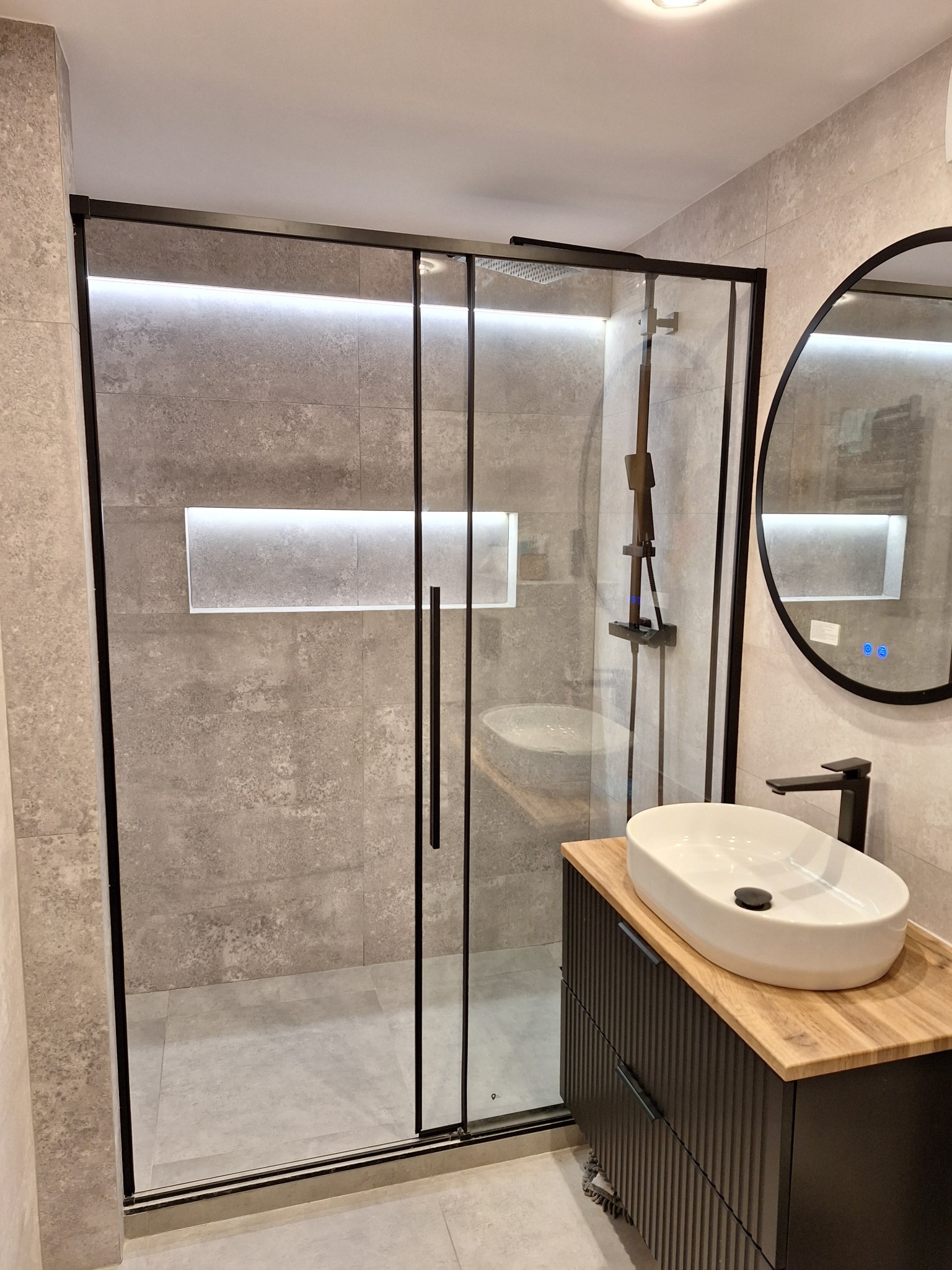 Nowoczesna łazienka z kabiną prysznicową typu walk-in z czarnymi profilami, umywalką nablatową na drewnianym blacie i okrągłym lustrem z podświetleniem LED.