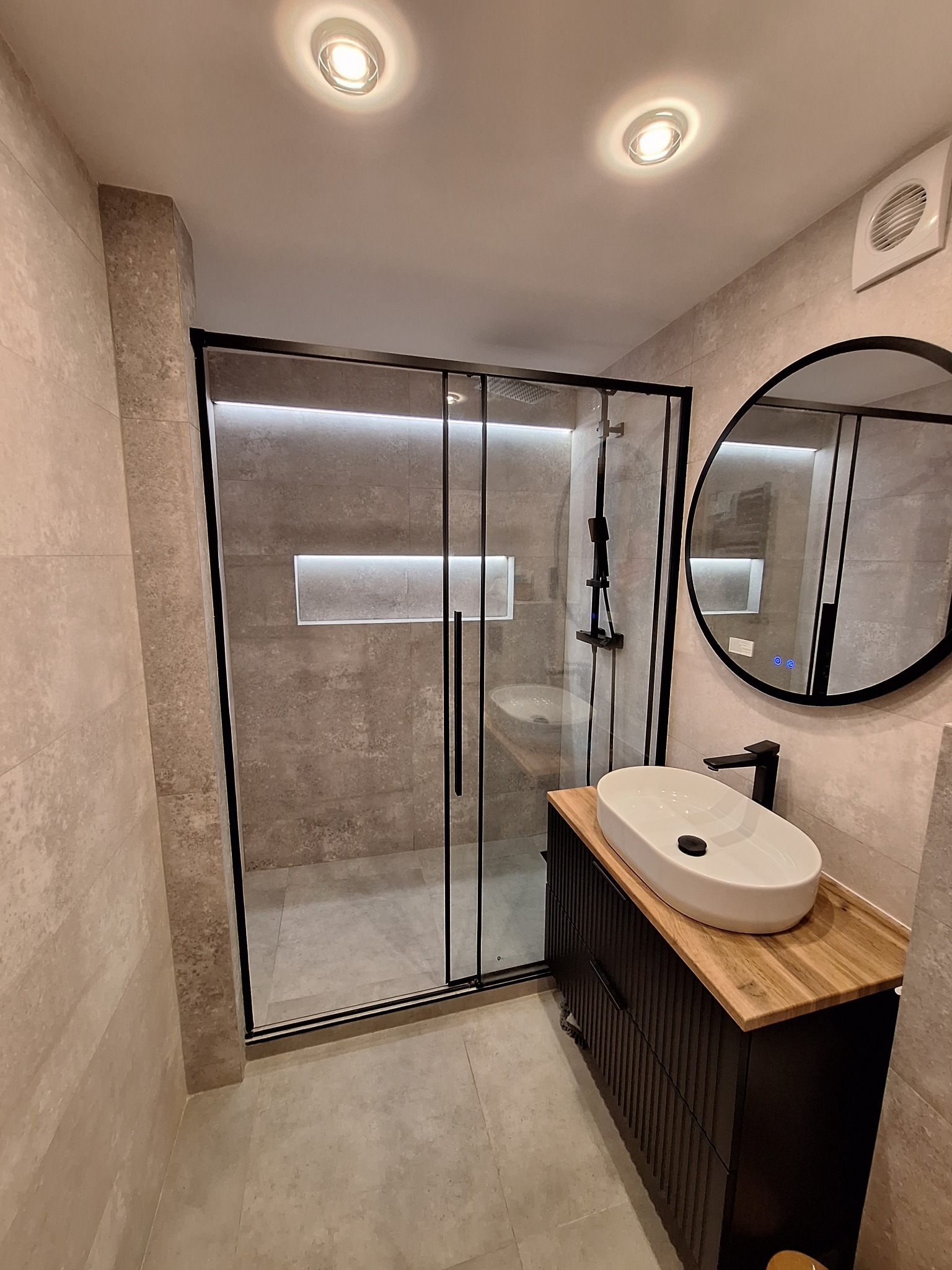 Nowoczesna łazienka z kabiną prysznicową typu walk-in z czarnymi profilami, owalną umywalką nablatową na szafce z drewnianym blatem i okrągłym lustrem z podświetleniem LED.