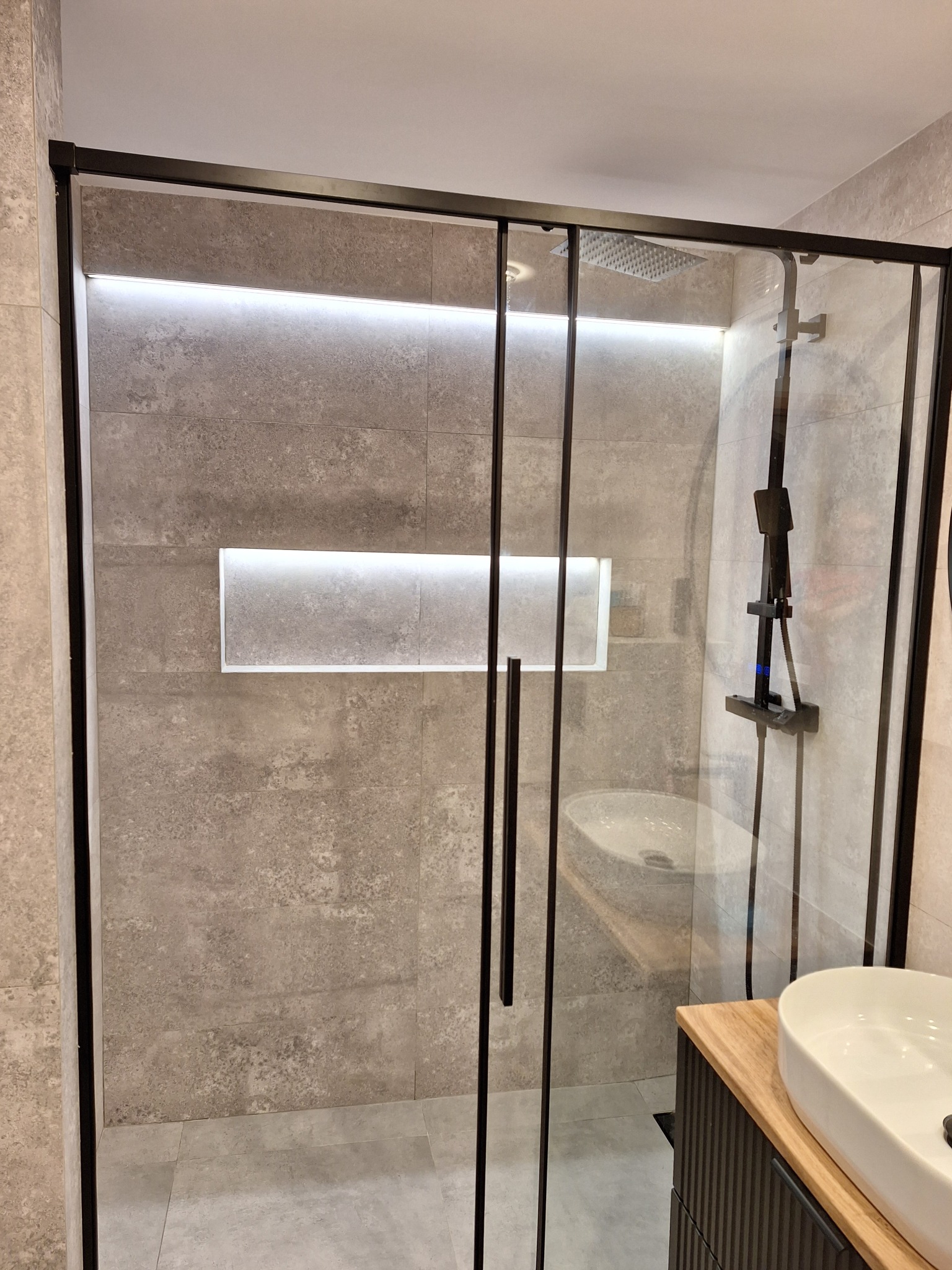 Nowoczesna kabina prysznicowa z czarnymi ramami i deszczownicą, w tle umywalka nablatowa na szafce z drewnianym blatem, podświetlana wnęka w ścianie wyłożonej szarymi płytkami.