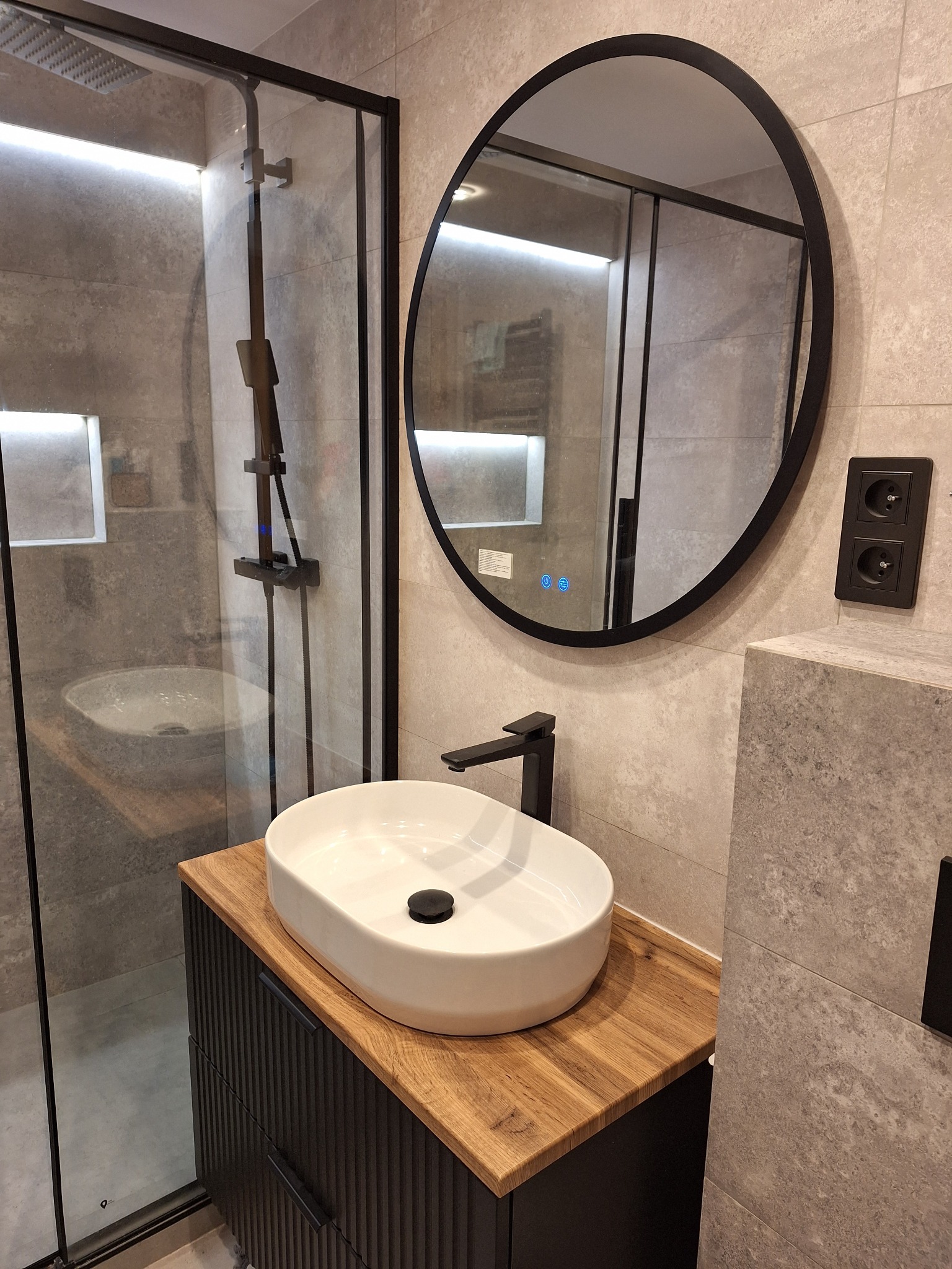 Nowoczesna łazienka z owalną białą umywalką nablatową na drewnianym blacie, czarna bateria, okrągłe lustro w czarnej ramie z podświetleniem LED, kabina prysznicowa z czarnymi profilami.