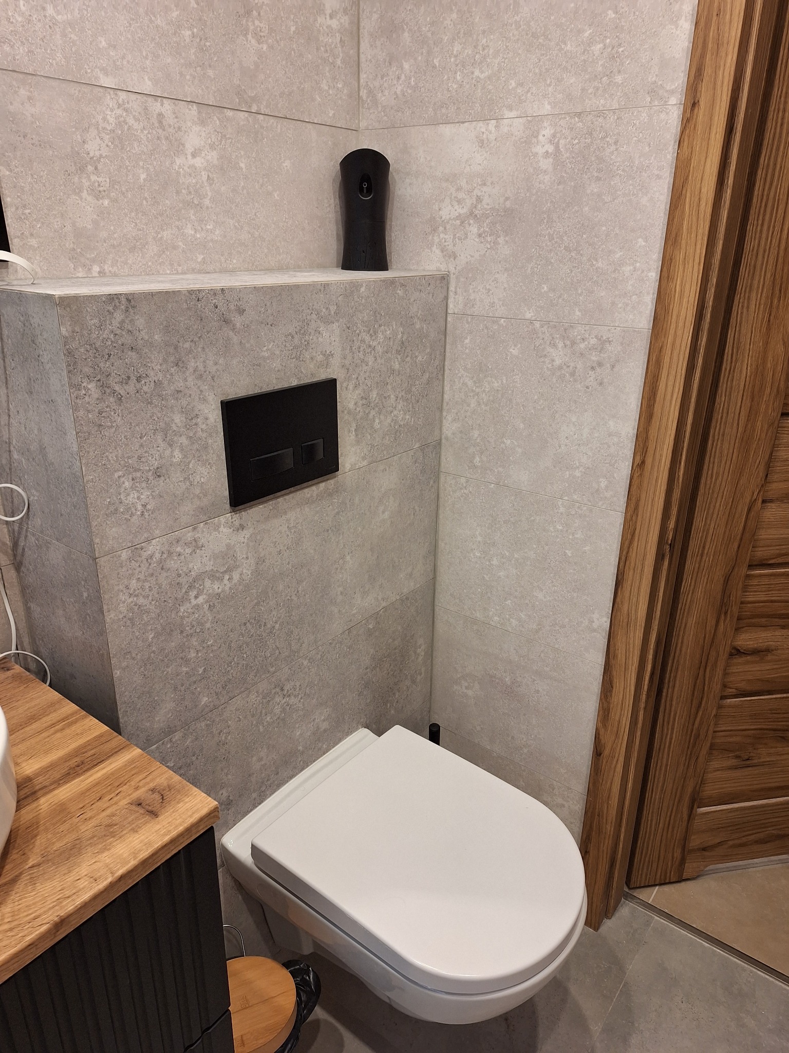 Nowoczesna toaleta podwieszana z czarnym przyciskiem spłukującym na tle płytek imitujących kamień, obok drewniane drzwi i fragment szafki umywalkowej z drewnianym blatem.