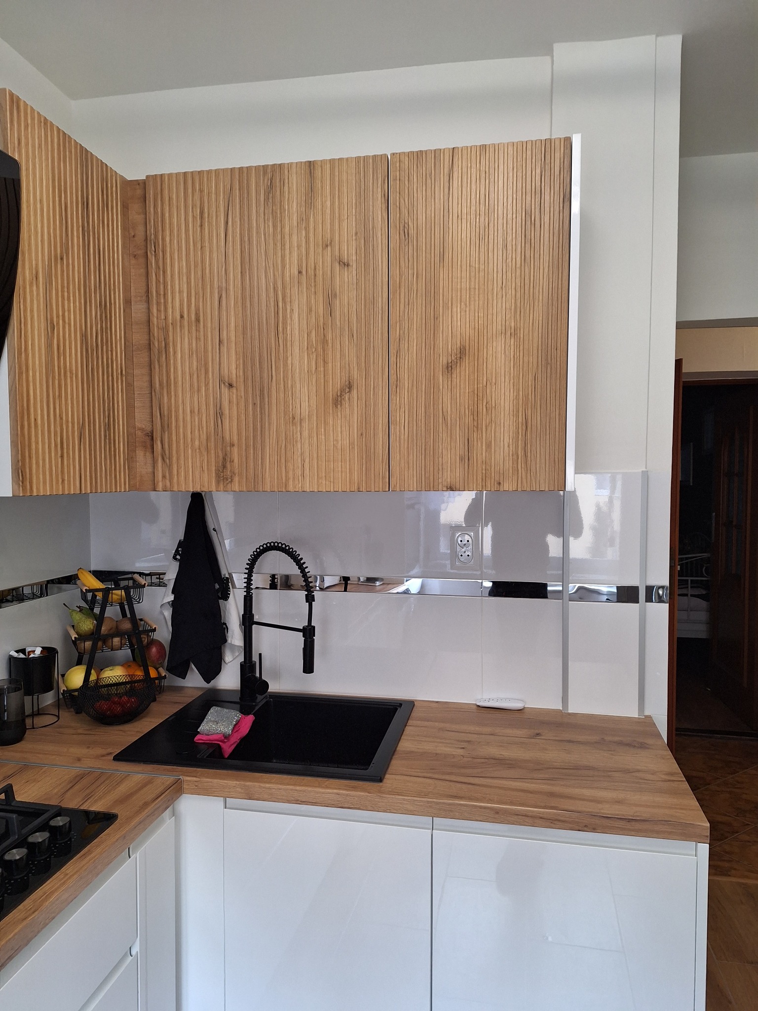 Nowoczesna kuchnia z drewnianymi szafkami górnymi o ryflowanej fakturze, białymi szafkami dolnymi z połyskiem, czarnym zlewem i baterią oraz blatem imitującym drewno. Na blacie widoczny koszyk...