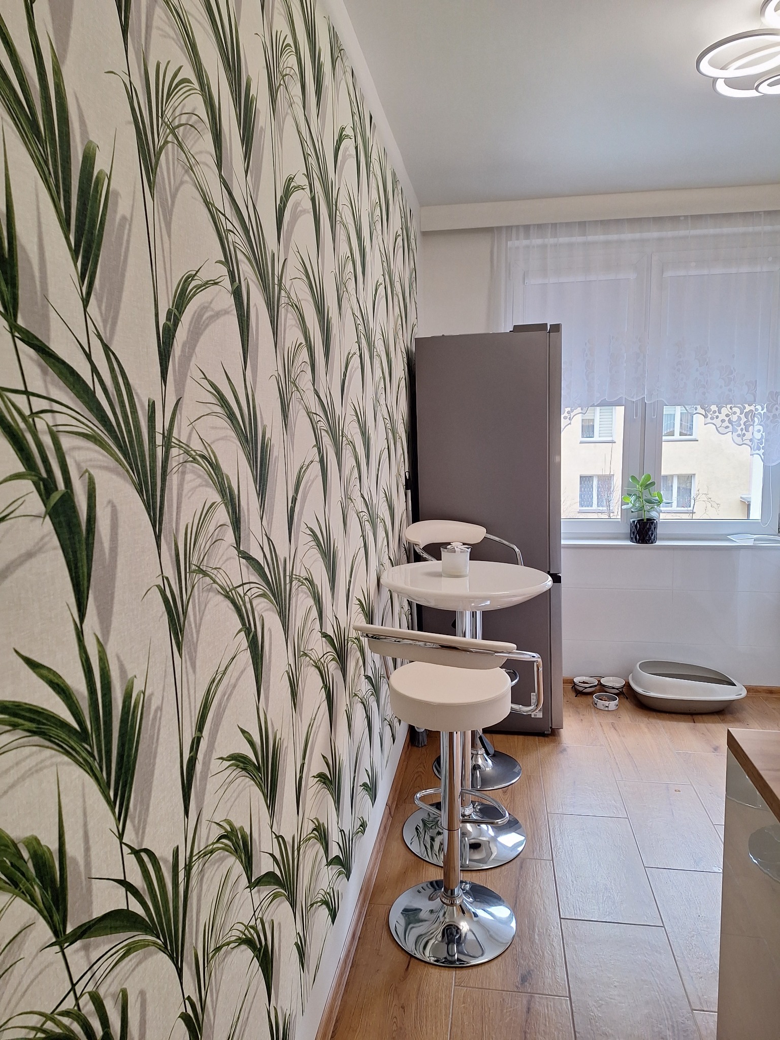 Białe krzesła barowe przy okrągłym stoliku w kuchni z tapetą w motywy roślinne, obok stoi szara lodówka, a na podłodze leżą miski dla zwierząt i kuweta.