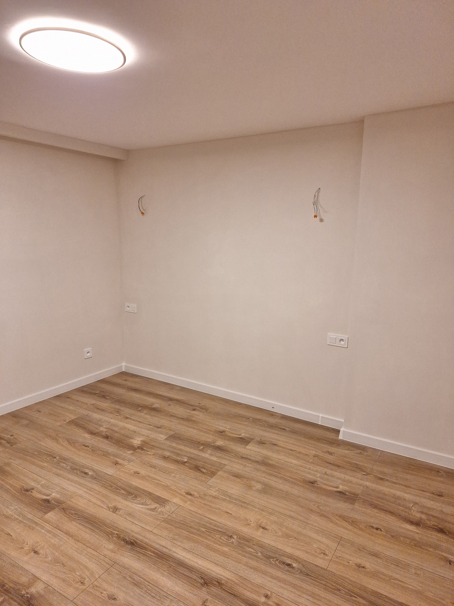 Pusty pokój po remoncie: nowe panele podłogowe w odcieniach brązu, świeżo pomalowane ściany w kolorze ecru, gniazdka elektryczne i wystające kable ze ściany. Widoczne białe listwy przypodłogowe...