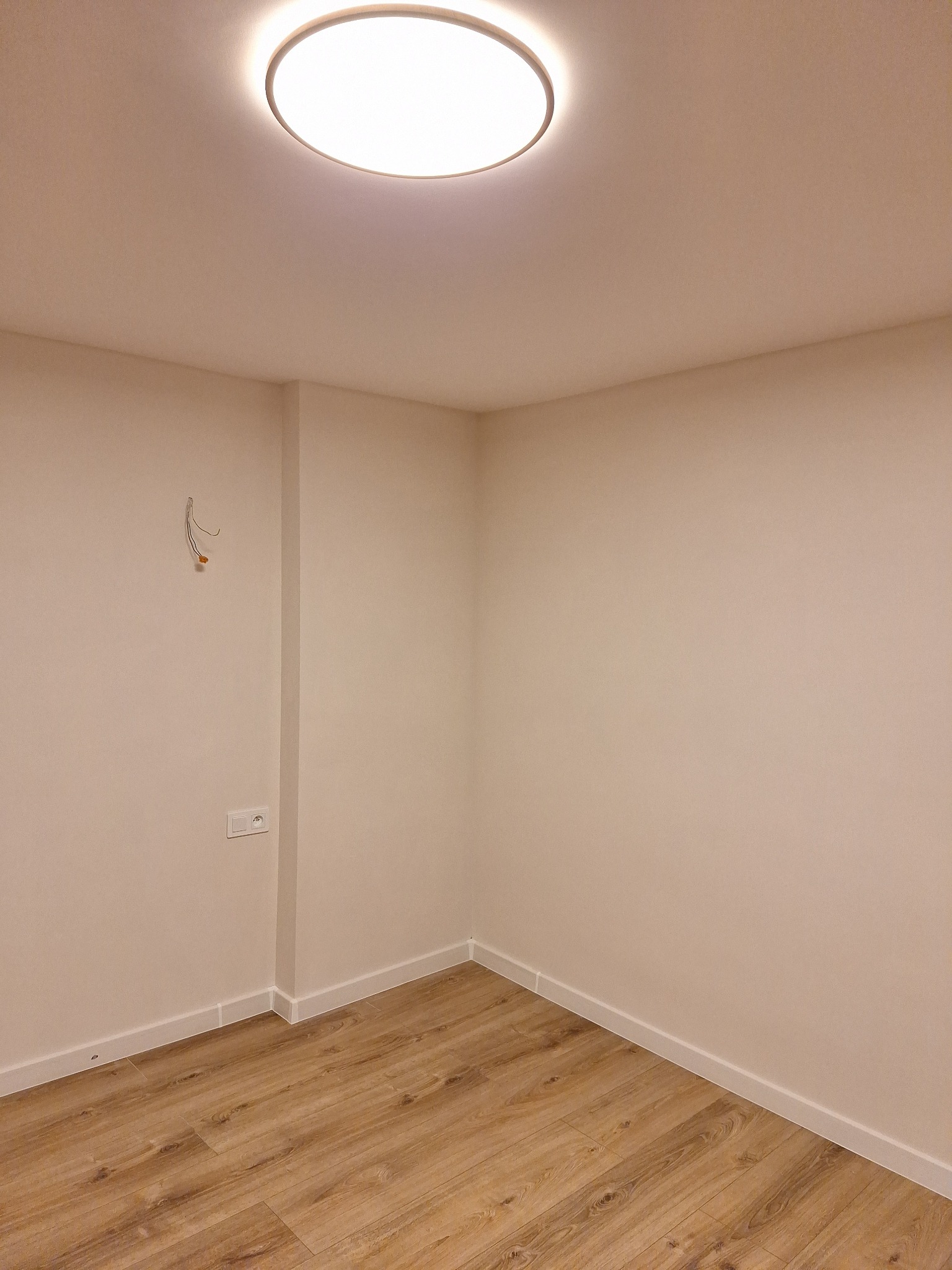 Pusty pokój po remoncie z panelami podłogowymi, pomalowanymi ścianami na jasny kolor i zamontowanym oświetleniem sufitowym. Widoczne wystające kable ze ściany oraz gniazdko elektryczne.