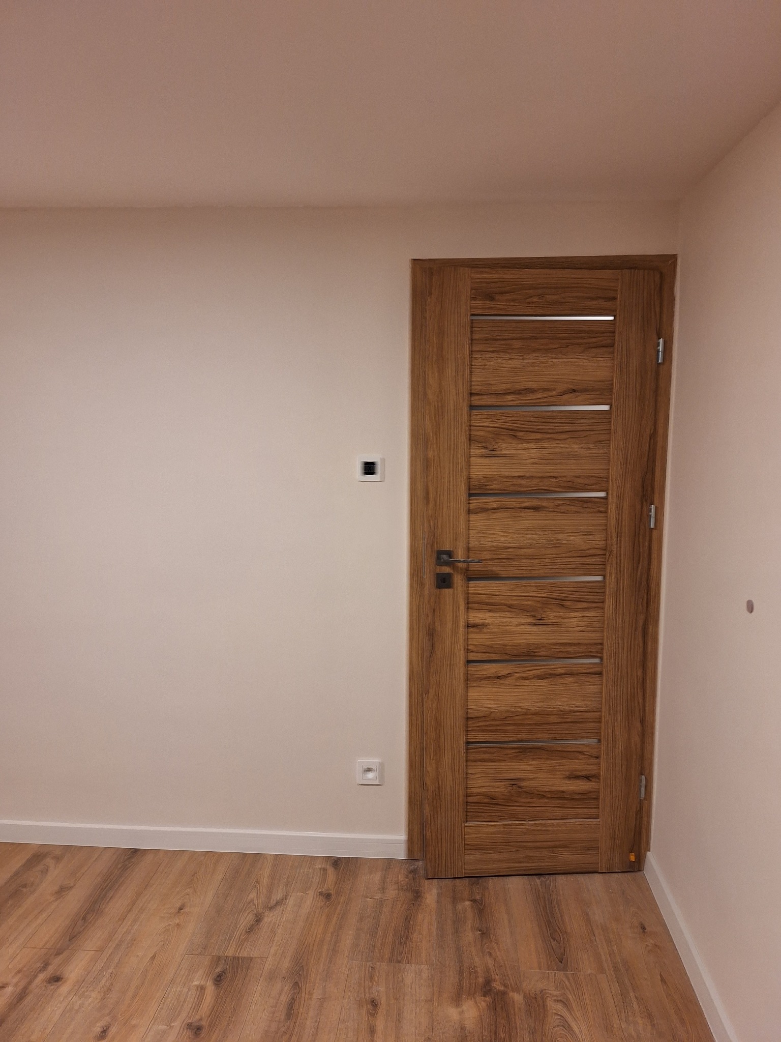 Wnętrze pokoju z drewnianą podłogą i drzwiami wewnętrznymi z poziomymi, metalowymi wstawkami, widoczny kontakt i włącznik na ścianie.