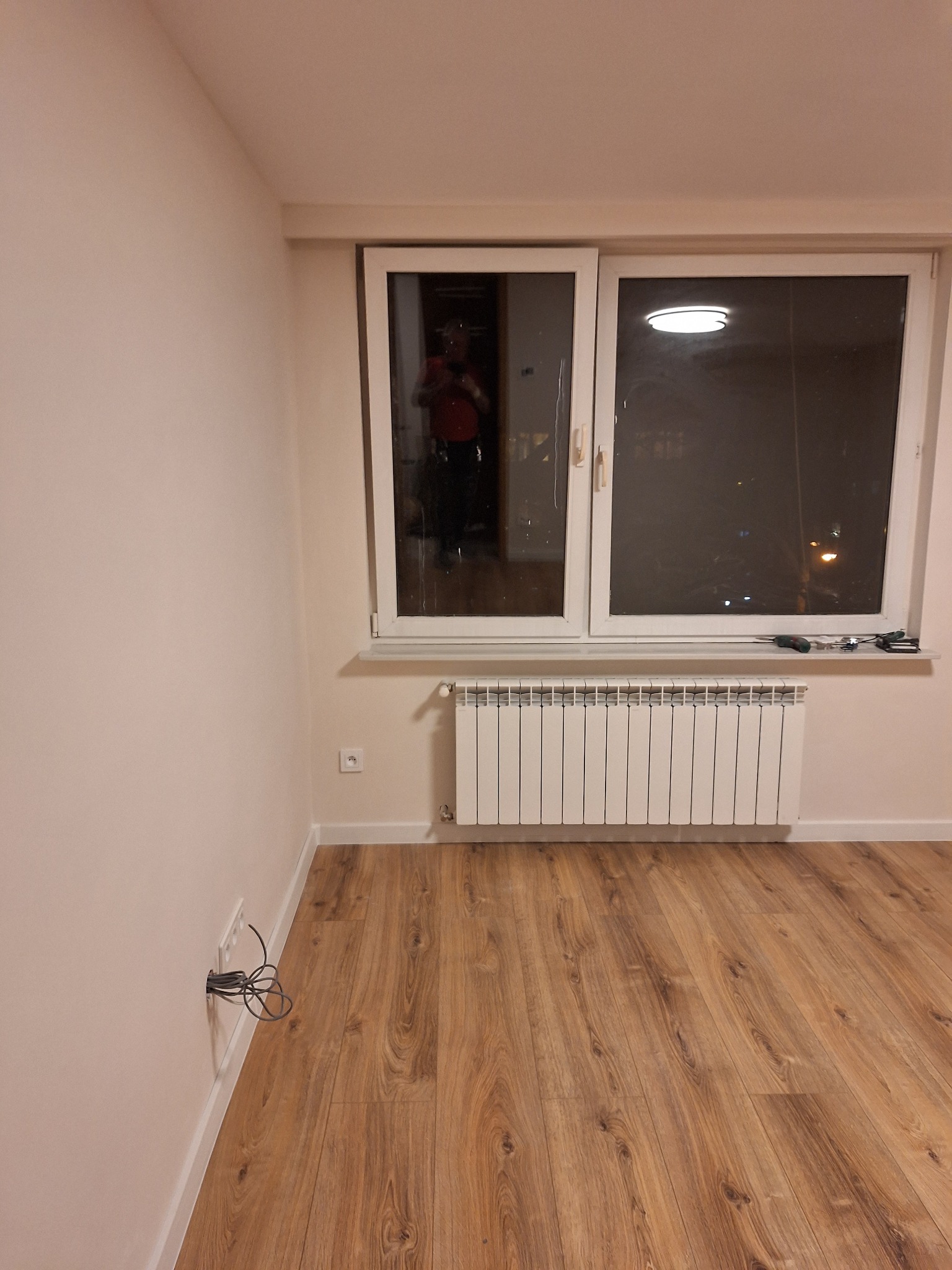 Pusty pokój po remoncie z drewnianą podłogą, białymi ścianami i grzejnikiem pod oknem. Na parapecie leżą narzędzia, a w oknie widać odbicie osoby robiącej zdjęcie. Ze ściany wystają kable.