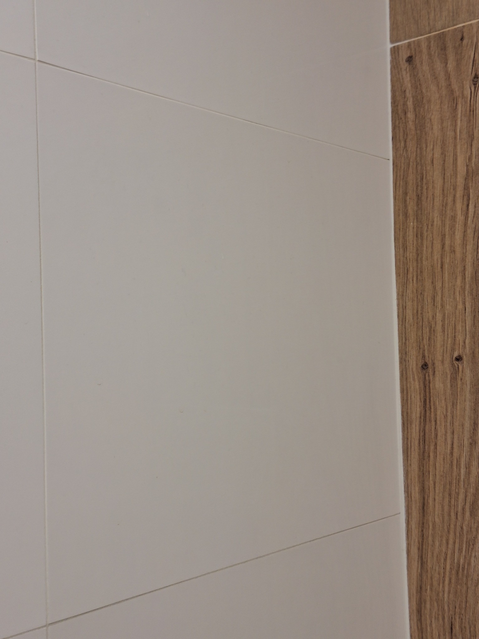 Fragment ściany wyłożonej jasnymi, kwadratowymi płytkami ceramicznymi, przylegającej do pionowego panelu z imitacją drewna z widocznymi słojami.