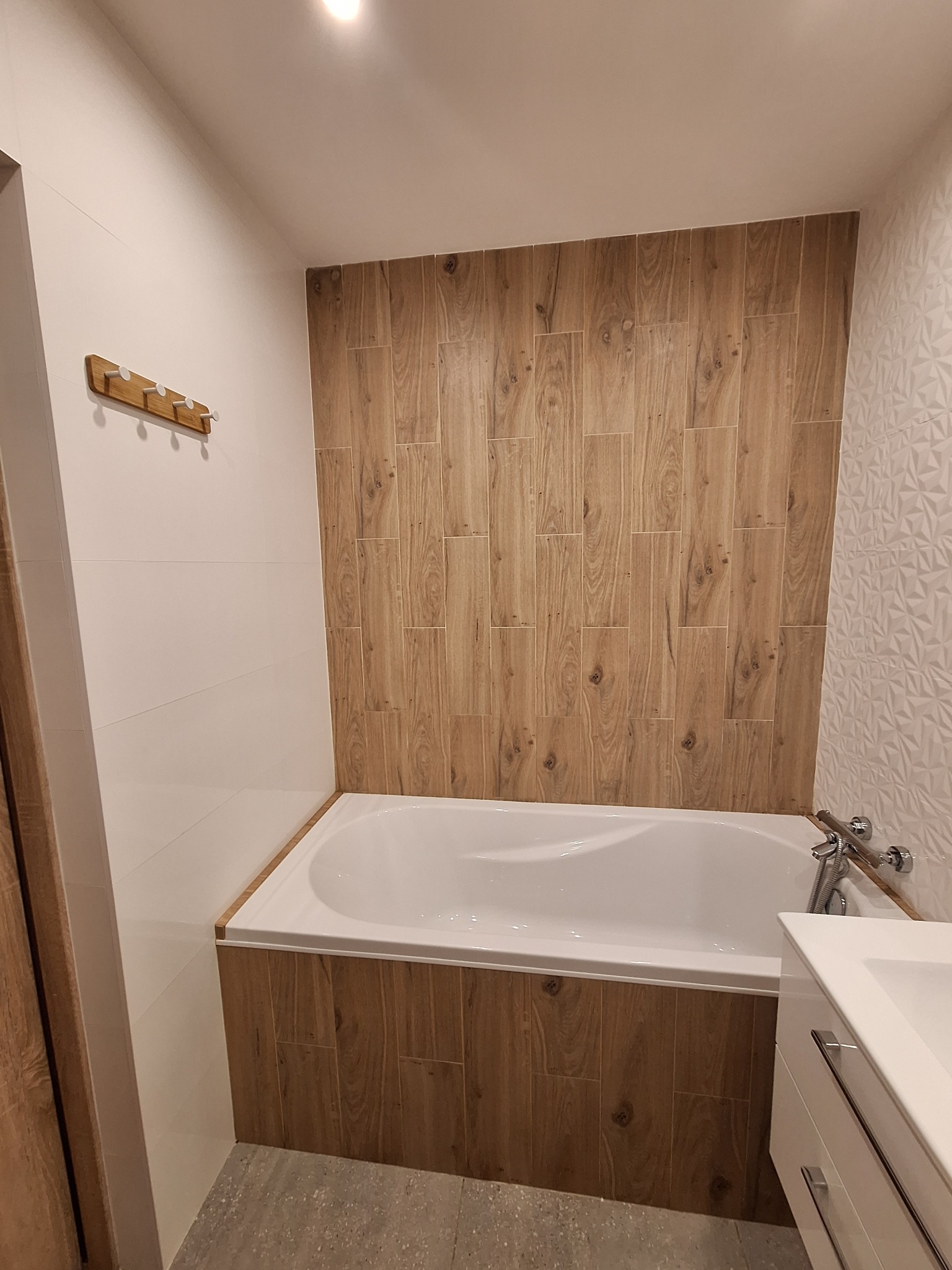 Nowoczesna łazienka z wanną obudowaną płytkami imitującymi drewno, białe płytki 3D na ścianie i drewniany wieszak na ręczniki.