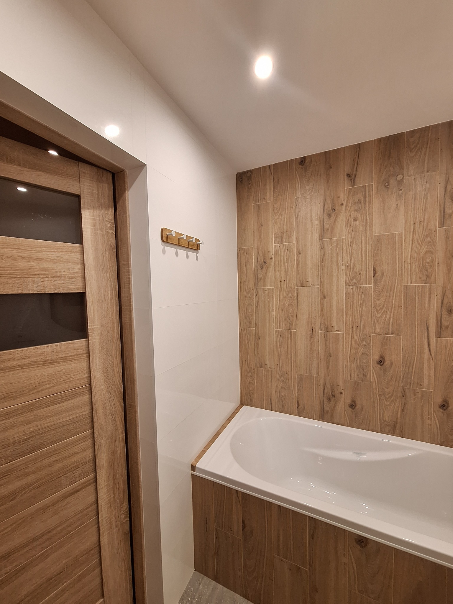Nowoczesna łazienka z wanną obudowaną płytkami imitującymi drewno, białe płytki na ścianie, drewniane drzwi z przeszkleniami i wieszak na ręczniki.