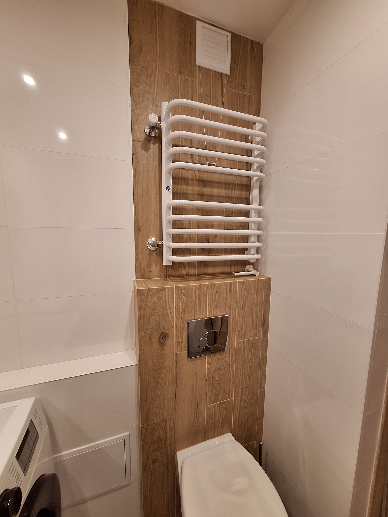 Nowoczesna łazienka z białymi płytkami i drewnopodobnymi panelami, grzejnik drabinkowy i częściowo widoczna pralka