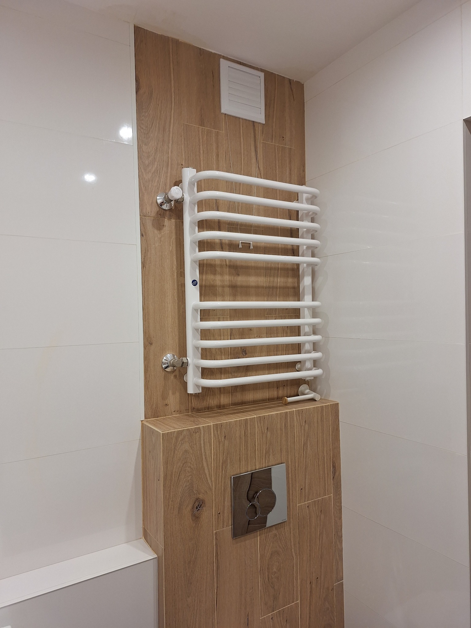 Biały, drabinkowy grzejnik łazienkowy na ścianie wyłożonej płytkami imitującymi drewno, obok białe, błyszczące płytki i przycisk spłuczki WC