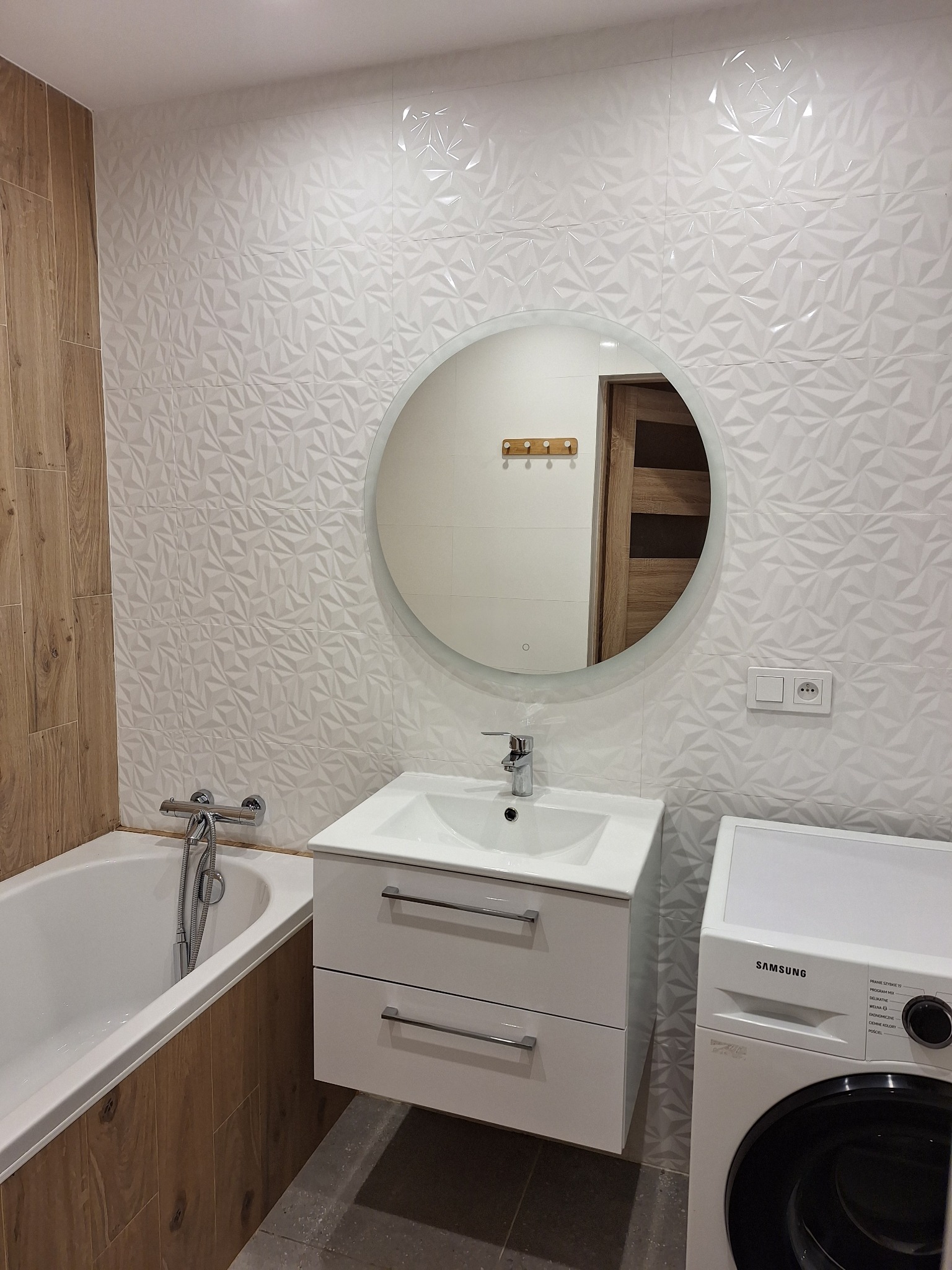 Nowoczesna łazienka z umywalką na szafce, okrągłym lustrem z podświetleniem LED, białą armaturą, pralką Samsung i ścianą wyłożoną trójwymiarowymi białymi płytkami.