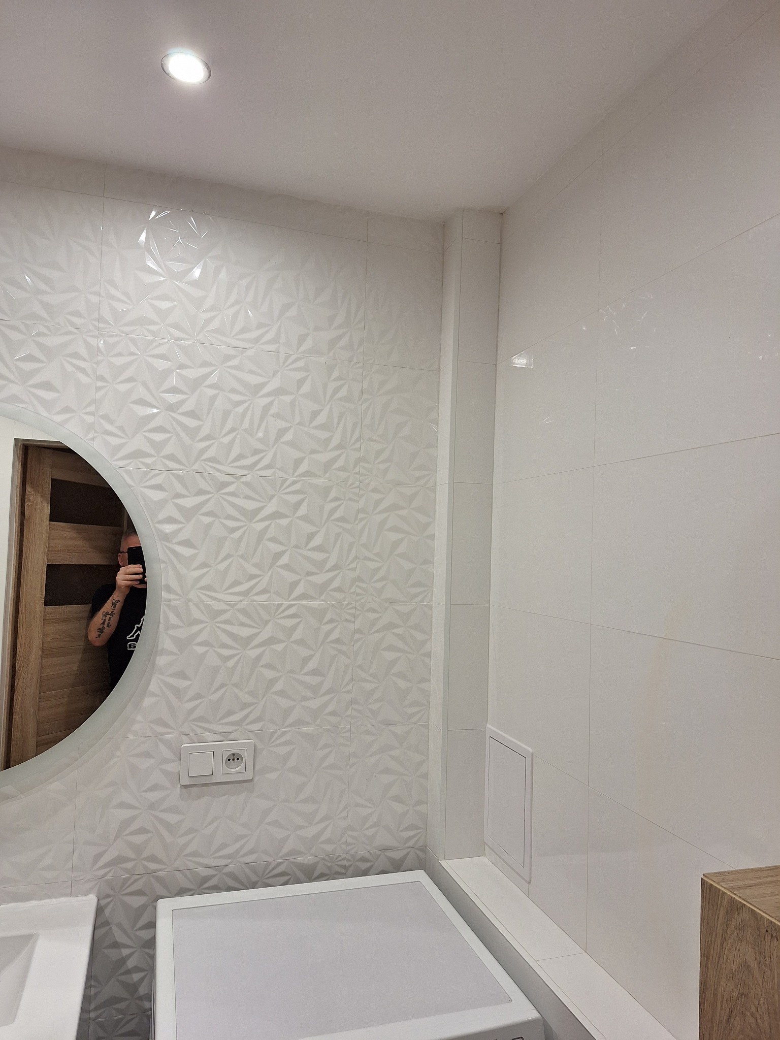 Biała łazienka z umywalką, pralką i okrągłym lustrem, ściany wyłożone płytkami 3D w geometryczne wzory, widoczne gniazdko elektryczne i rewizja.