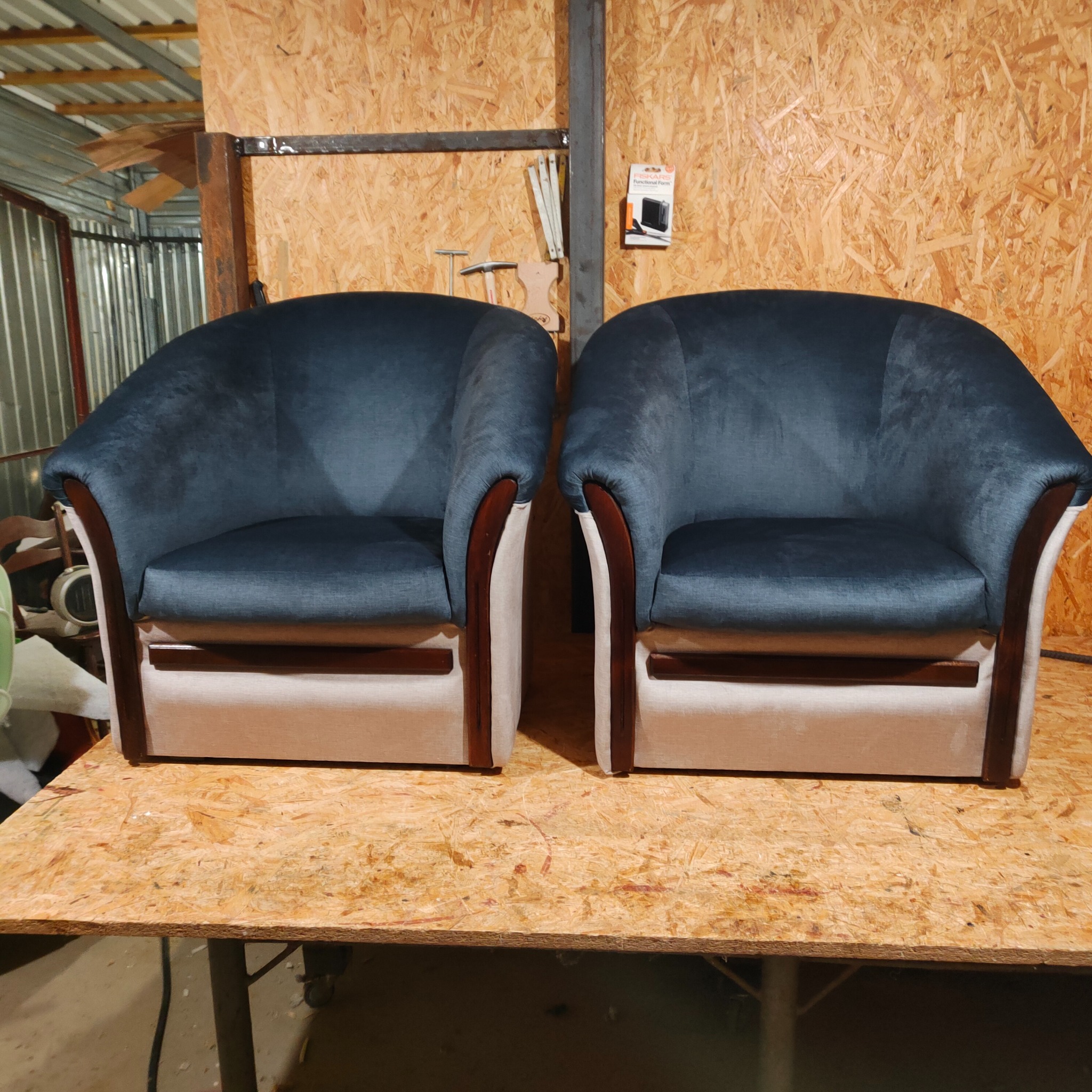 Dwa identyczne, odnowione fotele klubowe z ciemnoniebieską tapicerką, kremowymi bokami i mahoniowymi akcentami, stojące na roboczym stole z płyty OSB w warsztacie.