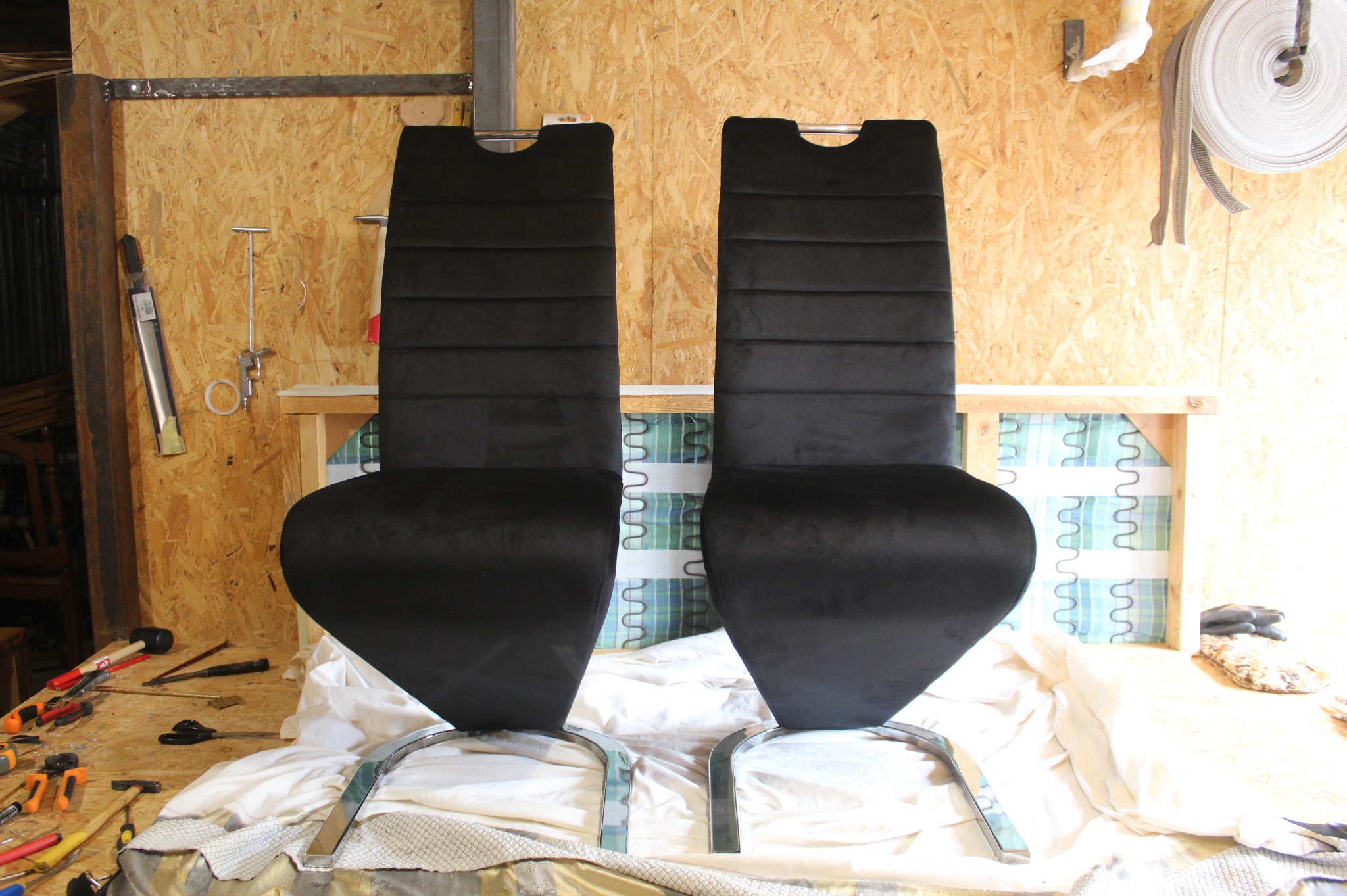 Dwa czarne, tapicerowane krzesła z chromowanymi nogami w warsztacie tapicerskim, widoczne narzędzia i materiały, w tle ściana z płyty OSB.