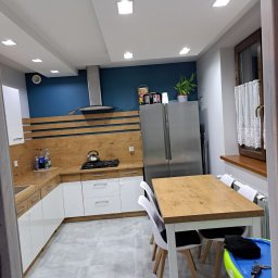 Usługi remontowe i wykończeniowe - Nowoczesna kuchnia z białymi szafkami, drewnianymi blatami i akcentem w postaci ciemnoniebieskiej ściany. Widoczny stół z krzesłami, stalowa lodówka side-by-side oraz okno z firanką.