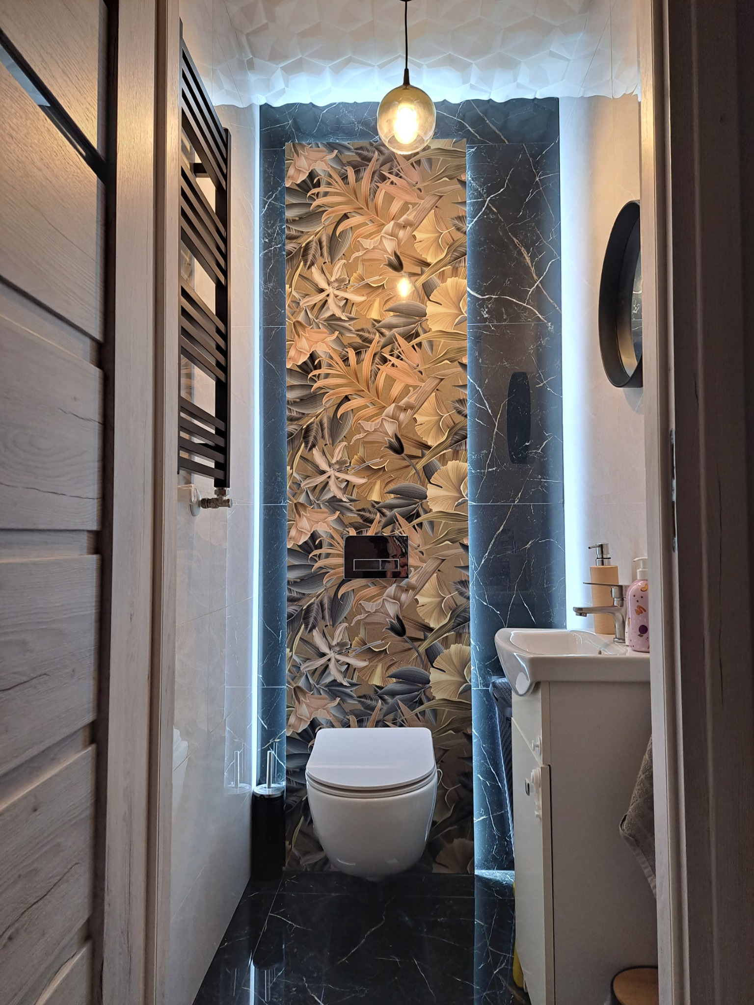 Wykończona łazienka z podświetlaną ścianą z motywem roślinnym, czarną marmurową podłogą i sufitem z geometryczną fakturą, oświetlona wiszącą lampą z żarówką Edison.
