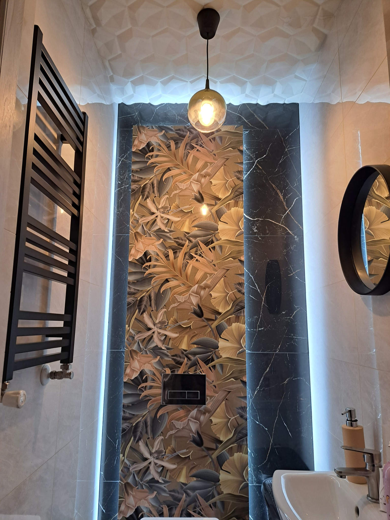Nowoczesna łazienka z czarnym grzejnikiem drabinkowym, panelem ściennym w kwiaty, marmurowymi płytkami podświetlonymi ledami, okrągłym lustrem i wiszącą lampą z żarówką Edison