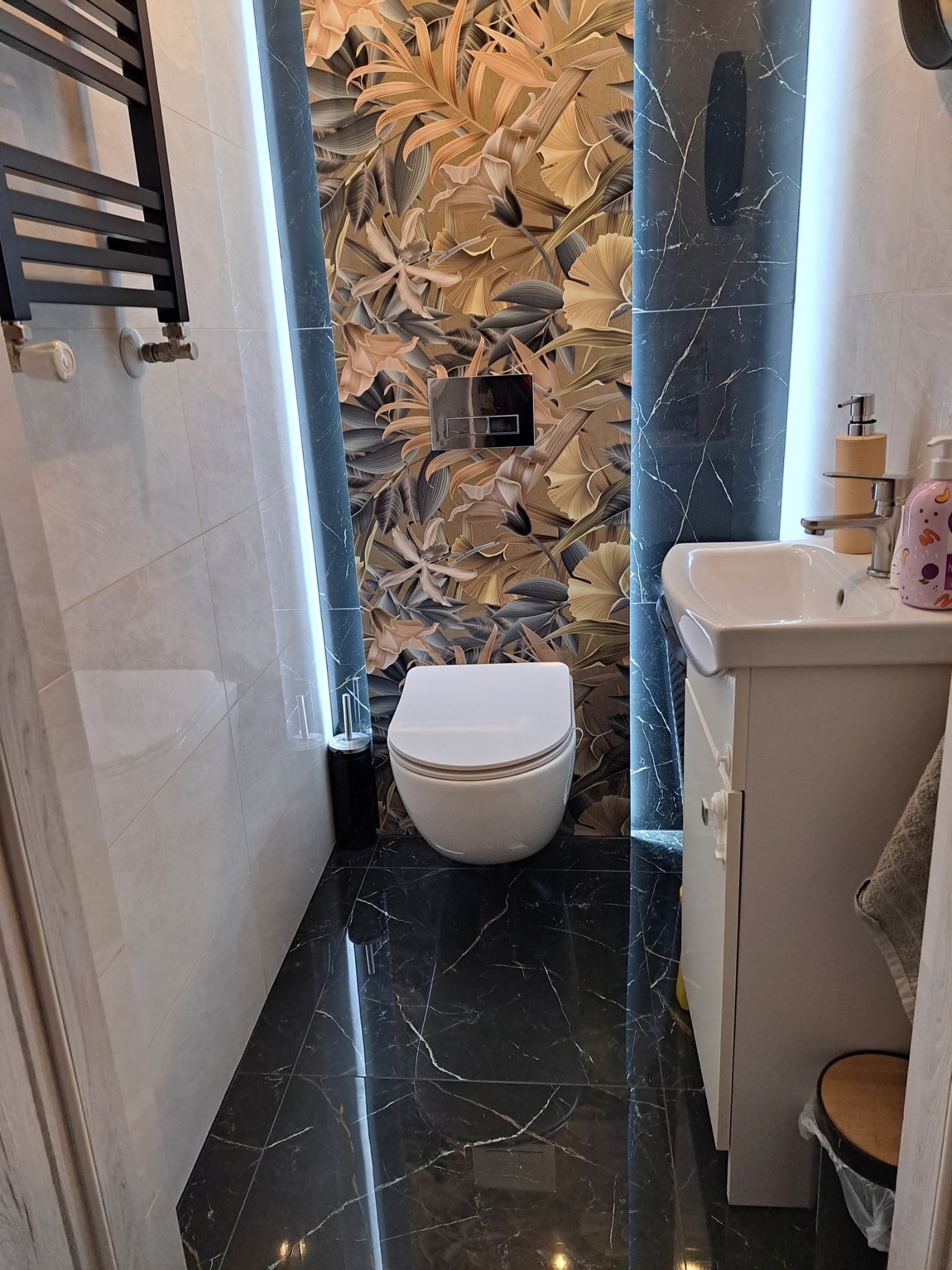 Nowoczesna łazienka z podświetlanymi panelami imitującymi marmur i tapetą w tropikalne liście, wisząca toaleta i czarna marmurowa podłoga z białymi żyłami.