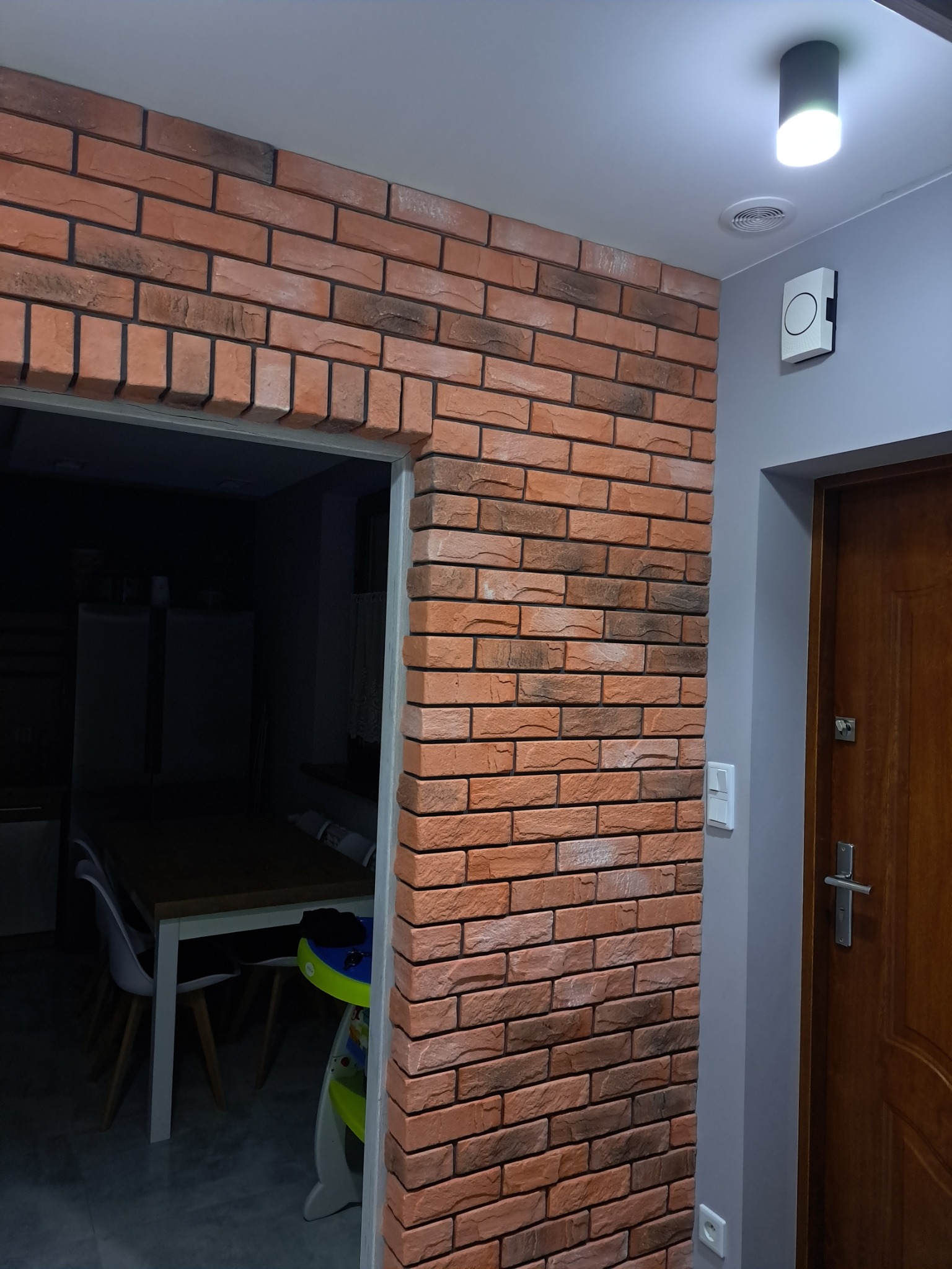 Ściana wyłożona dekoracyjną cegłą w odcieniach czerwieni i brązu, przylegająca do futryny drzwiowej prowadzącej do ciemnego pomieszczenia z widocznym stołem i krzesłami, obok drewniane drzwi...