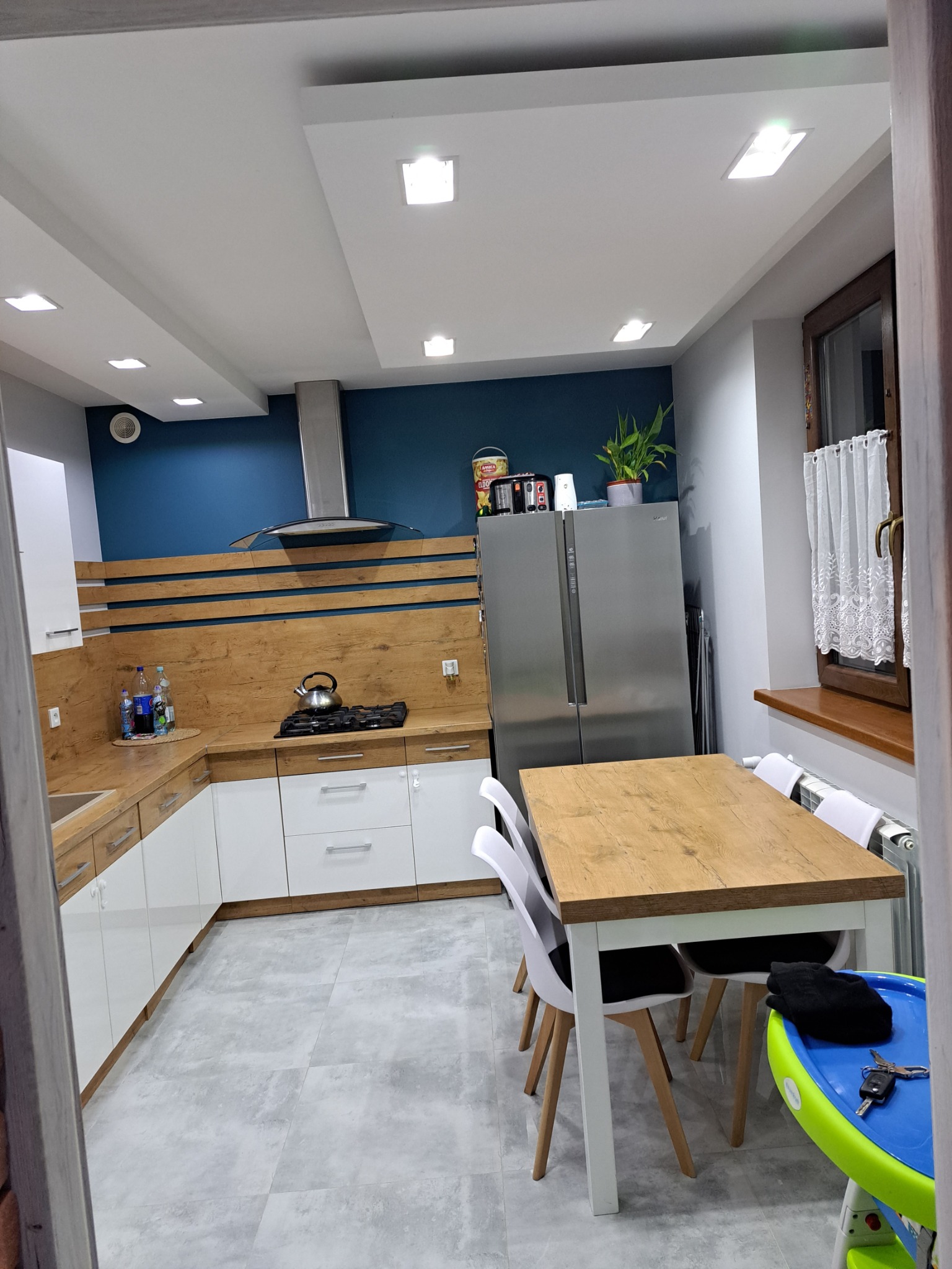 Nowoczesna kuchnia z białymi szafkami, drewnianymi blatami i akcentem w postaci ciemnoniebieskiej ściany. Widoczny stół z krzesłami, stalowa lodówka side-by-side oraz okno z firanką.